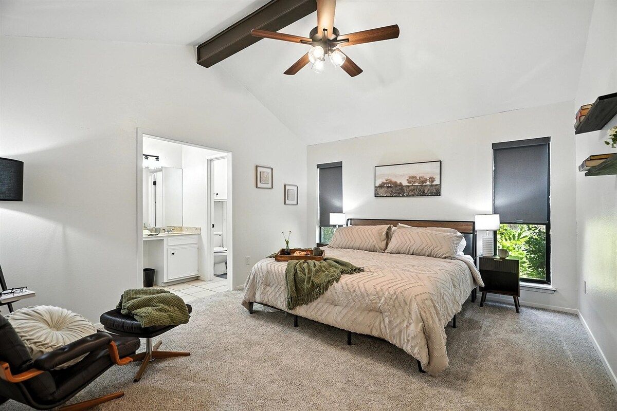 A bedroom with a bed, a chair, a bedside table, a ceiling fan, a window, and a door leading to another room.