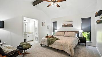 A bedroom with a bed, a chair, a bedside table, a ceiling fan, a window, and a door leading to another room.