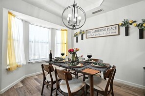 Un comedor con una mesa dispuesta para seis personas, rodeada de sillas, una araña y un letrero en la pared.