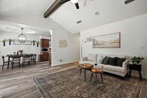 Amplia sala de estar con sofá, mesa de centro y un tablero de ajedrez. Hay un comedor con mesa y sillas, y una cocina al fondo.