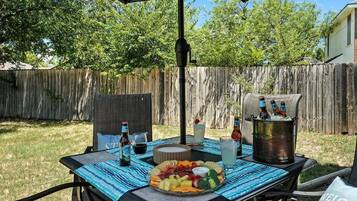 An outdoor patio with a table set for a meal, including a fruit platter, drinks, and a cooler.