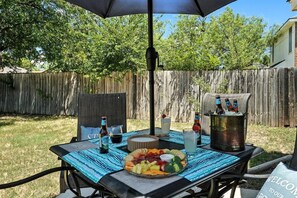 Une terrasse extérieure avec une table dressée pour un repas, comprenant un assortiment de fruits, des boissons et une glacière.
