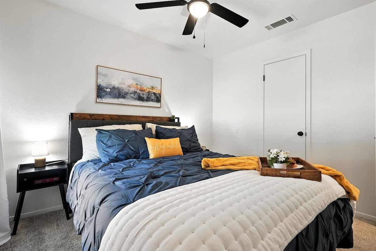Een slaapkamer met een bed, een nachtkastje, een plafondventilator, een deur en een kunstwerk aan de muur.