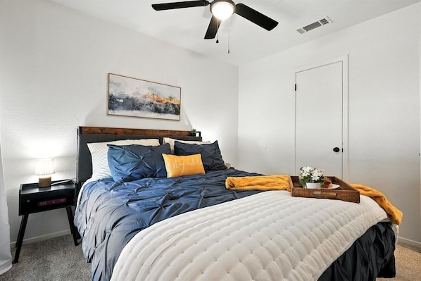 Un dormitorio con cama, mesita de noche, ventilador de techo, una puerta y una obra de arte en la pared.