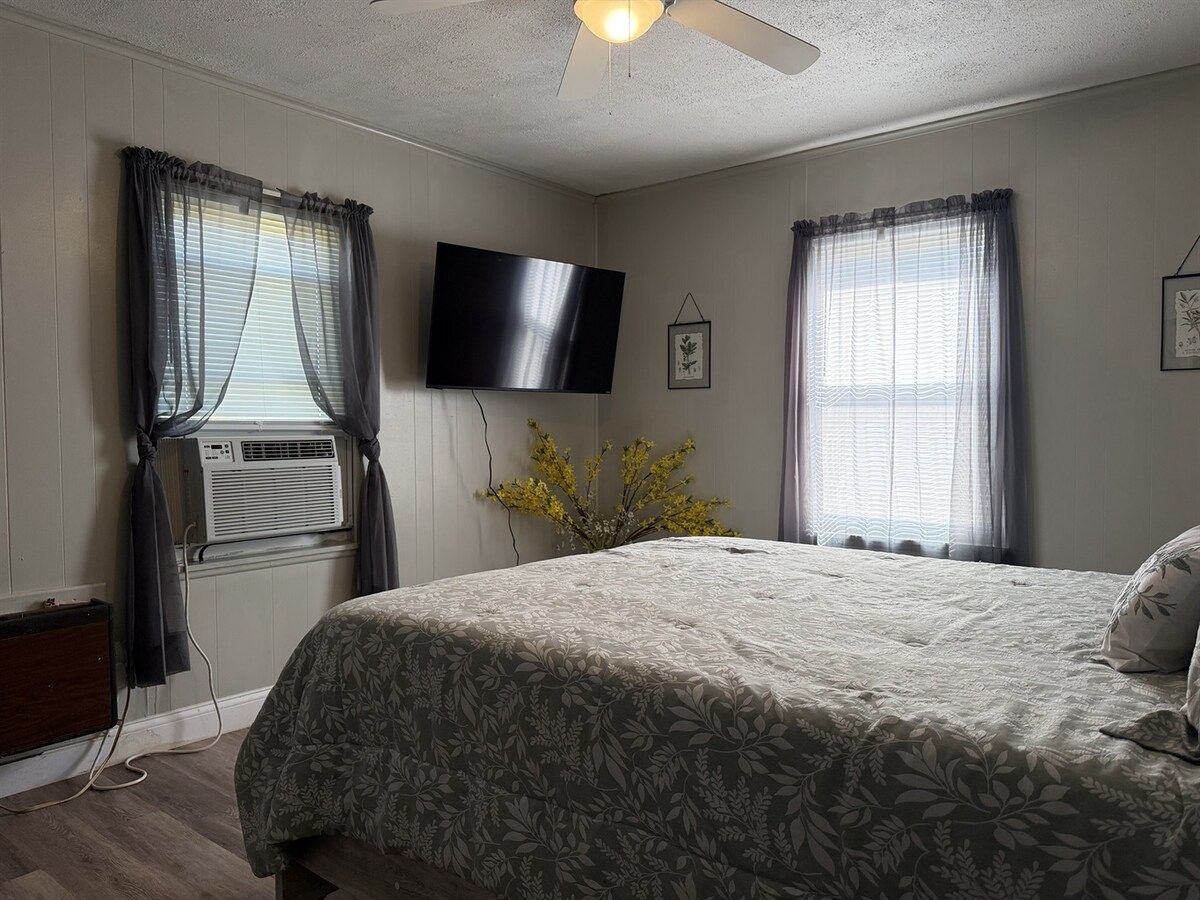 A bedroom with a bed, a flat-screen TV, two windows with curtains, an air conditioning unit, and a ceiling fan.