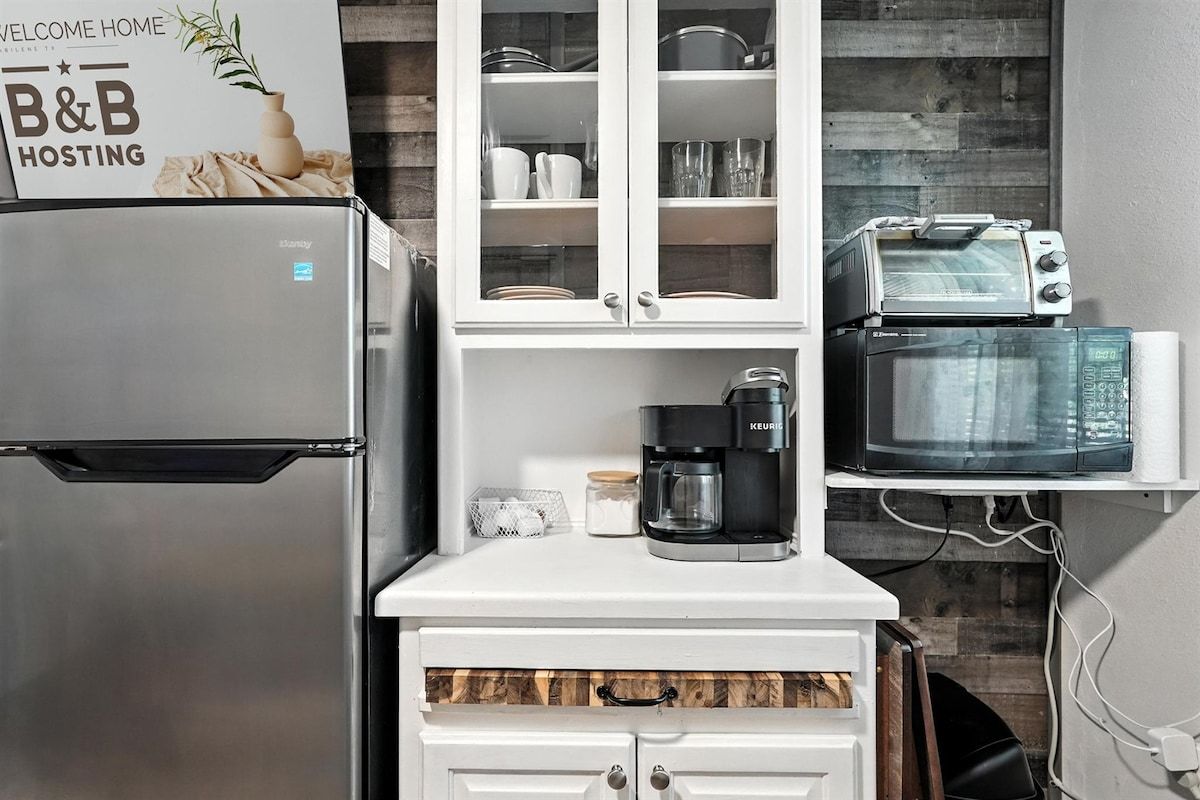 A compact kitchen with a stainless steel refrigerator, a coffee maker, a microwave, and a toaster. There is a cabinet with glass doors above the countertop.