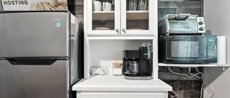 A compact kitchen with a stainless steel refrigerator, a coffee maker, a microwave, and a toaster. There is a cabinet with glass doors above the countertop.