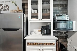 Une cuisine compacte équipée d’un réfrigérateur en acier inoxydable, d’une machine à café, d’un four à micro-ondes et d’un grille-pain. Un meuble avec des portes en verre se trouve au-dessus du plan de travail.
