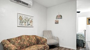 A living room with a floral sofa, a light-colored armchair, and a wall-mounted air conditioner. - Mockingbird Lane – Cozy Rustic Retreat (Abilene)