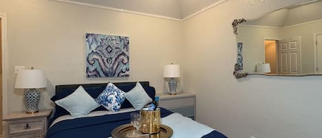 Ein ordentlich eingerichtetes Schlafzimmer mit einem Bett, zwei Nachttischen, einer Nachttischlampe, einem Tablett mit Tasse und Buch, einem Wandspiegel und einem Bild an der Wand.