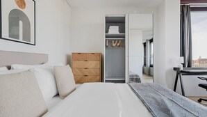 Une chambre d’hôtel moderne, équipée d’un lit, d’une commode en bois, d’un placard avec des vêtements suspendus, et offrant une vue sur la ville par la fenêtre.
