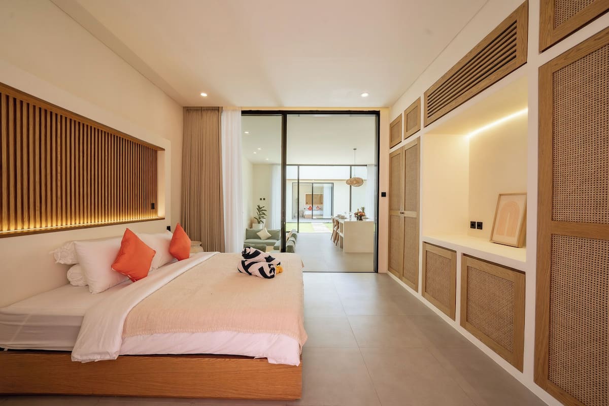 A modern bedroom with a large bed, a wooden headboard, and a view of a dining area through a glass door.