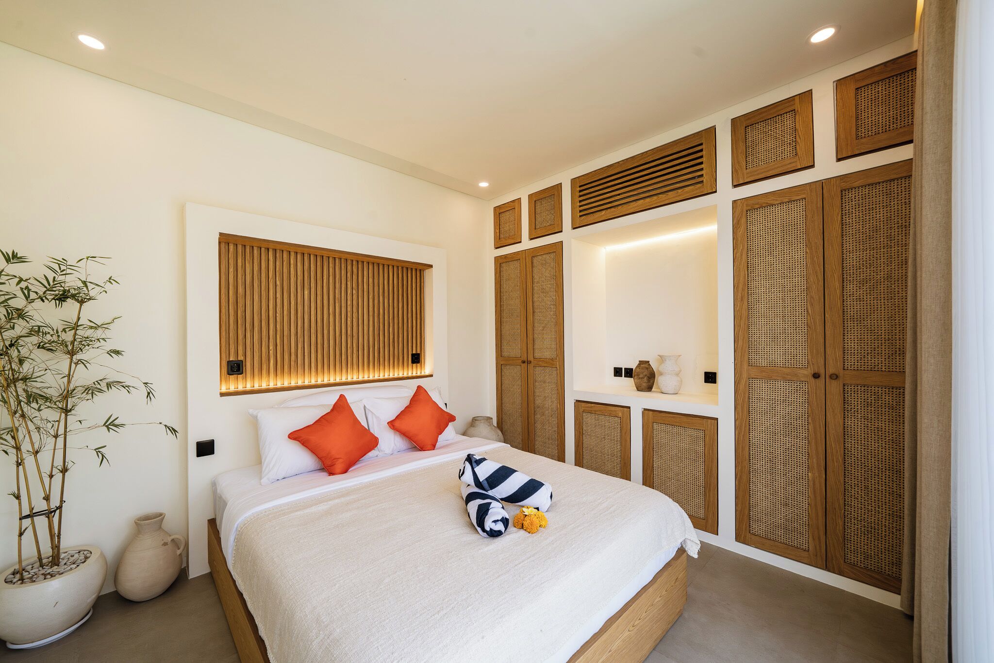 A modern hotel room with a large bed, a potted plant, and a wooden wardrobe with woven panels.