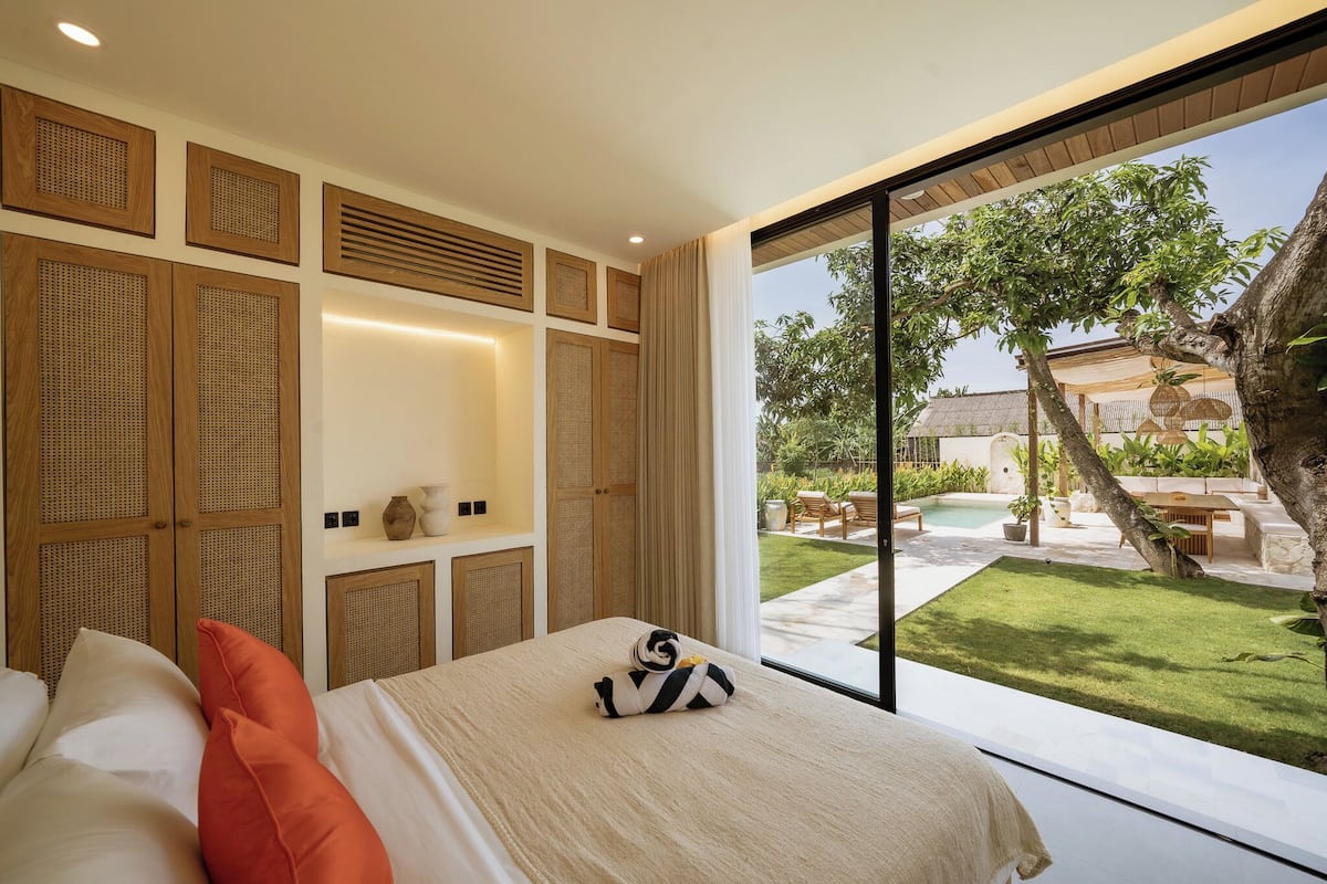 A bedroom with a large window, a bed with pillows, a bedside table, and a view of an outdoor swimming pool and garden.