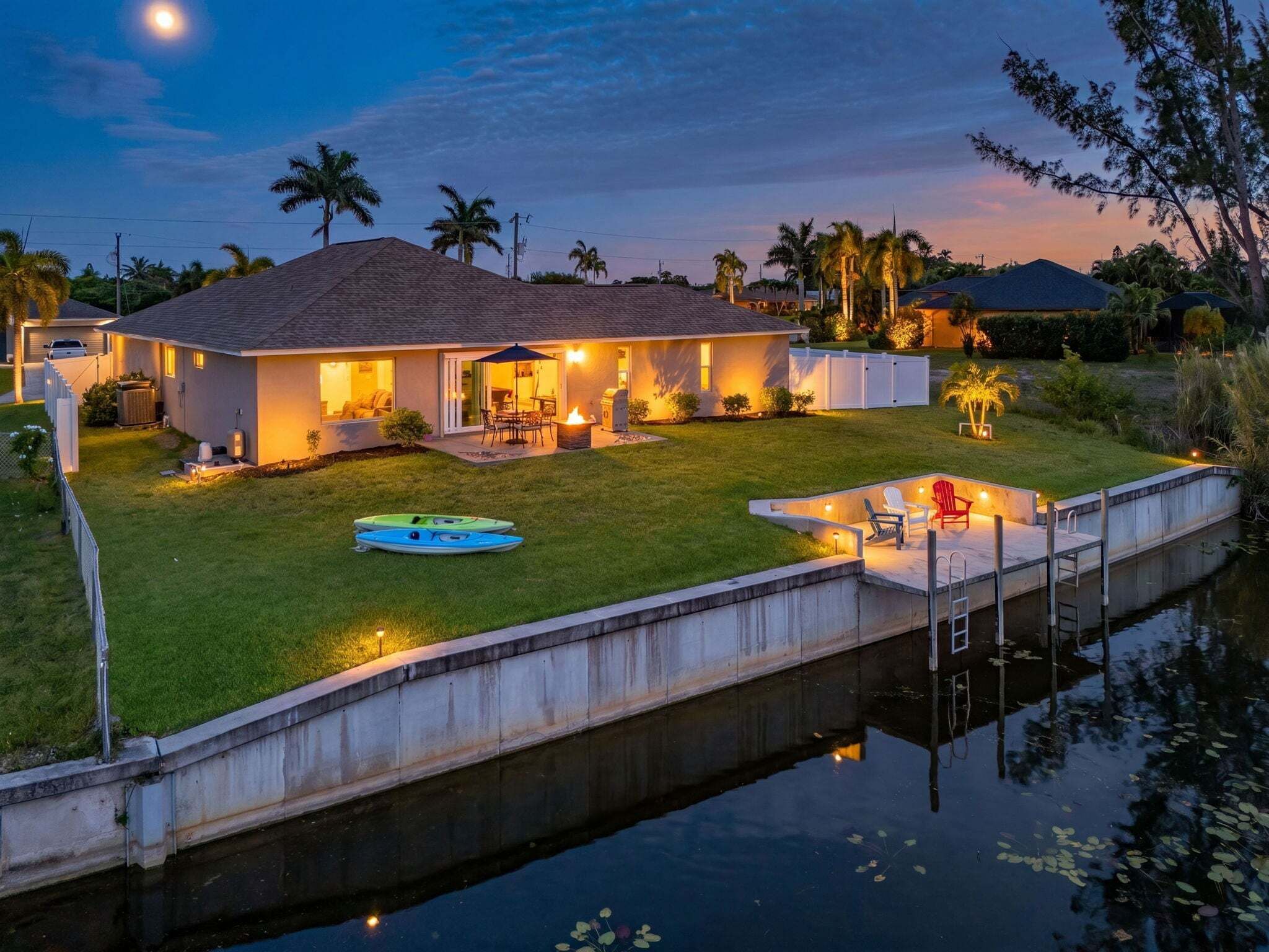 A house with a dock extending into a calm body of water, a well-maintained lawn, and a patio area with outdoor furniture.