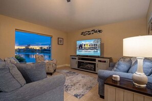 A living room with a large window, a flat-screen TV, a sofa, a lamp, and a decorative wall piece. - Waterfront Bliss with Pool & Spa – Your Perfect Escape Awaits - Sunset Cove - Roelens (Cape Coral)