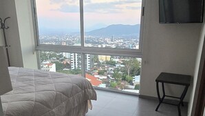 Quarto com cama, mesinha, televisão e uma grande janela com vista para a cidade.
