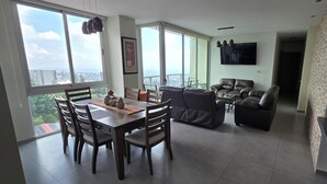 Uma sala de estar ampla, com uma mesa de jantar, dois sofás e uma TV fixada na parede.