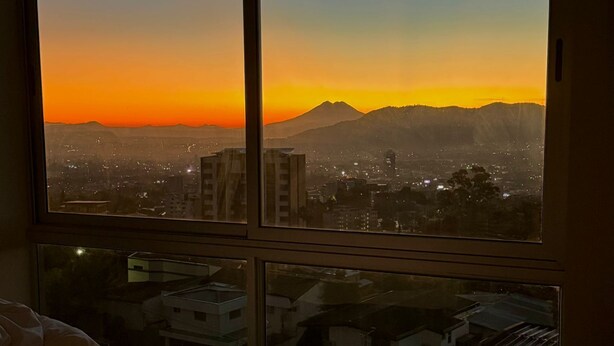 Quarto com uma grande janela com vista para a paisagem urbana ao pôr do sol.
