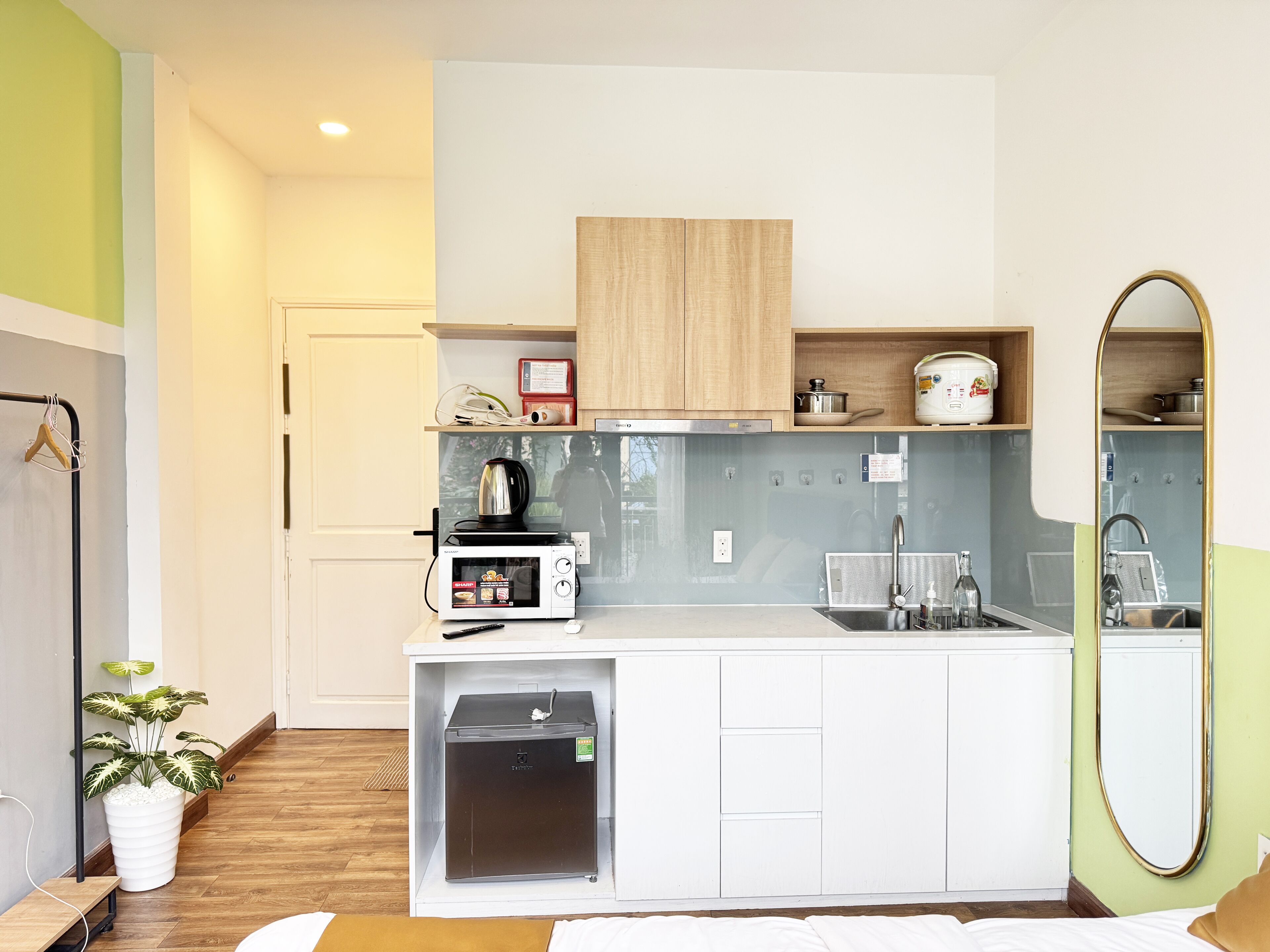 Une cuisine compacte équipée d’un four à micro-ondes, d’une bouilloire et d’un petit réfrigérateur. On y trouve un meuble en bois au-dessus du plan de travail et un miroir sur le mur.