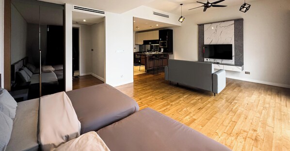 A modern living room with a sofa, a TV mounted on the wall, and a kitchen in the background. - Anggun Residence KLCC by Airhost (Kuala Lumpur)
