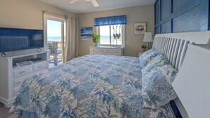 Ein Schlafzimmer mit Bett, Fernseher, einem Fenster mit Vorhängen, einer Pflanze und Meerblick.