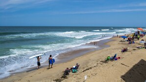 Ein Strand mit Menschen, einige laufen herum, andere tragen Surfbretter und entspannen auf Liegestühlen unter Sonnenschirmen.