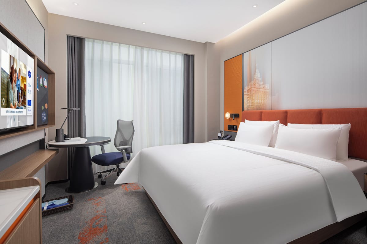 Ein Hotelzimmer mit einem großen Bett, einem Schreibtisch mit Stuhl, einem Fernseher und einem Fenster mit Vorhängen.