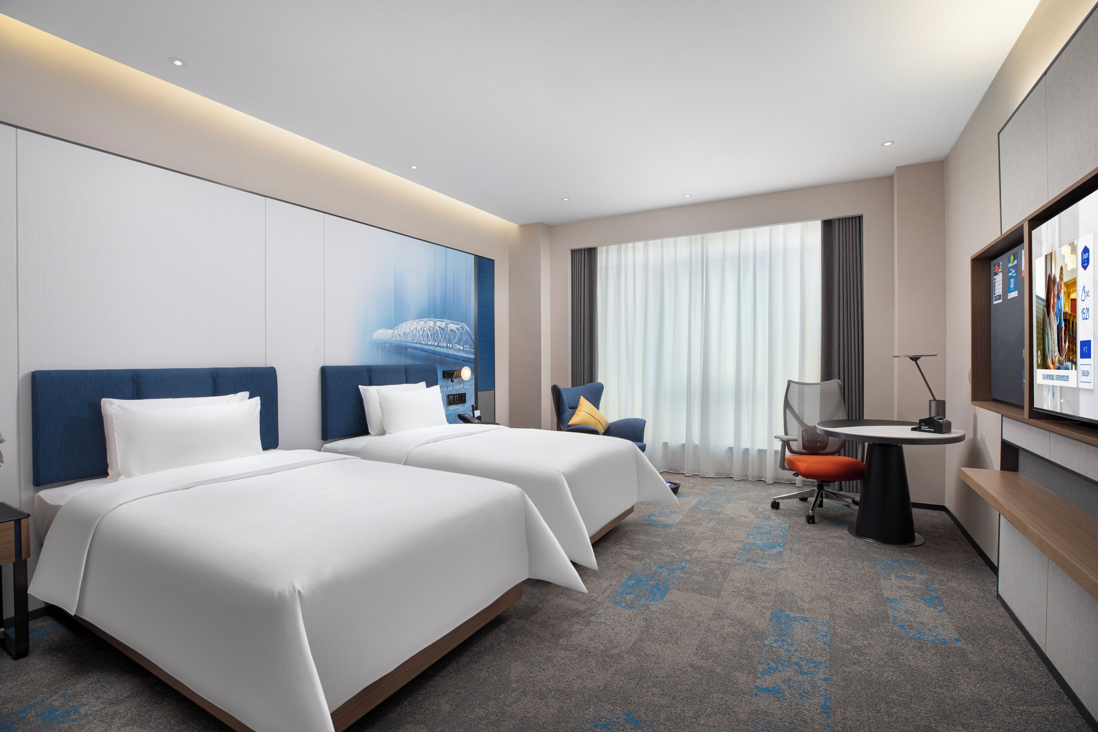 A hotel room with two beds, a desk, a chair, a TV, and a large window with curtains.