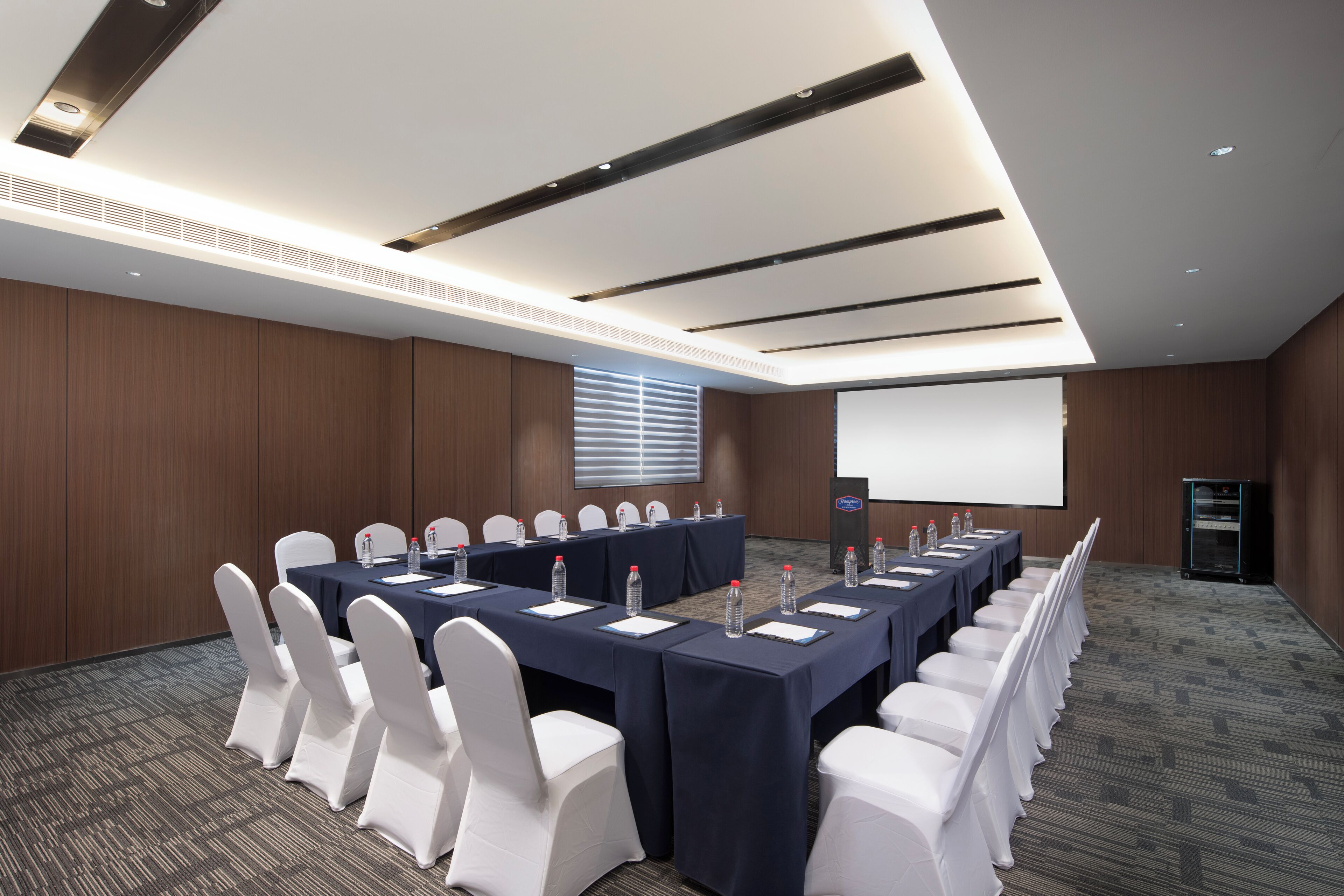 A conference room with a long table, white chairs, and a projector screen.