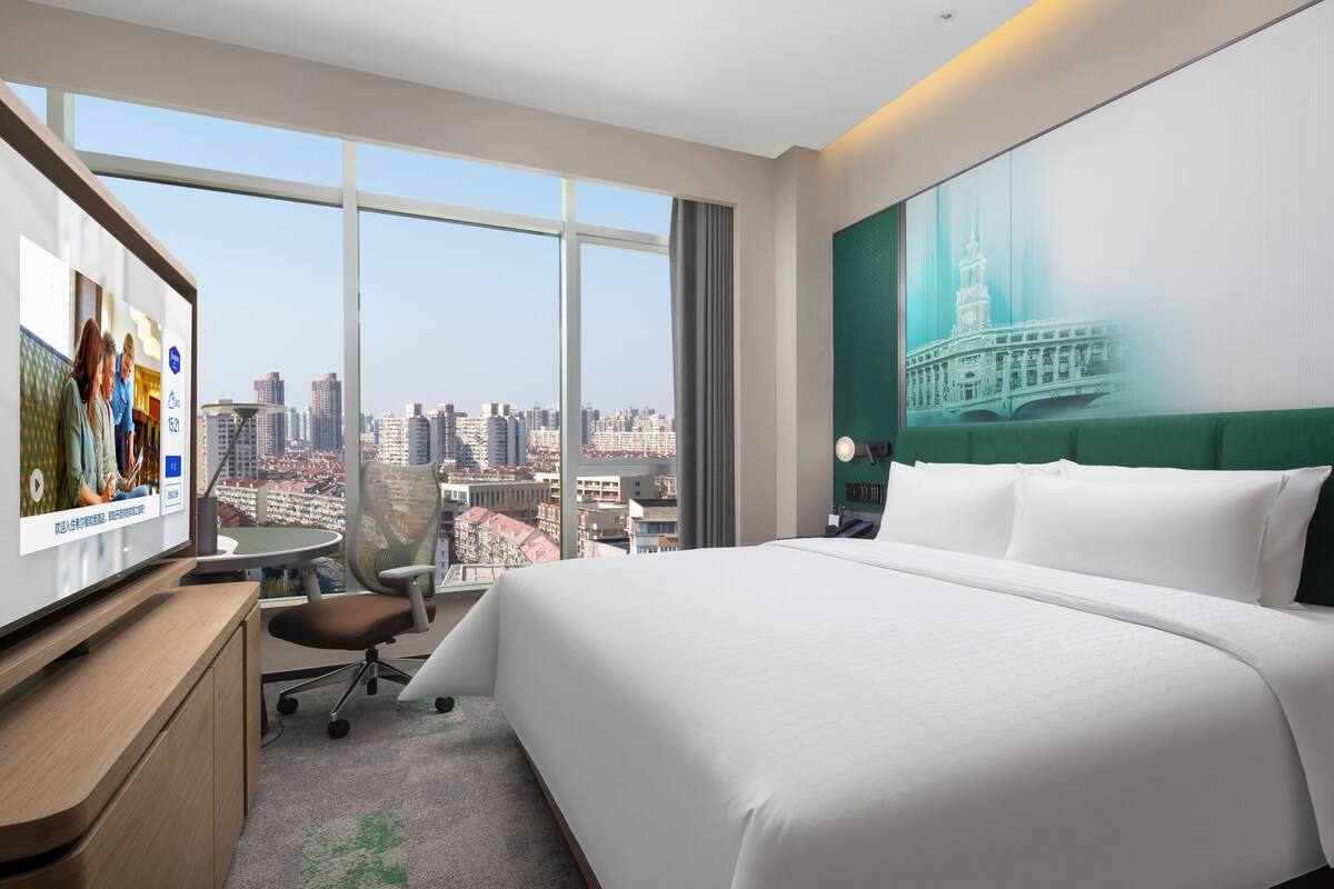 Ein Hotelzimmer mit einem großen Bett, einem Schreibtisch, einem Sessel und einem Fernseher, der eine Stadtansicht zeigt.
