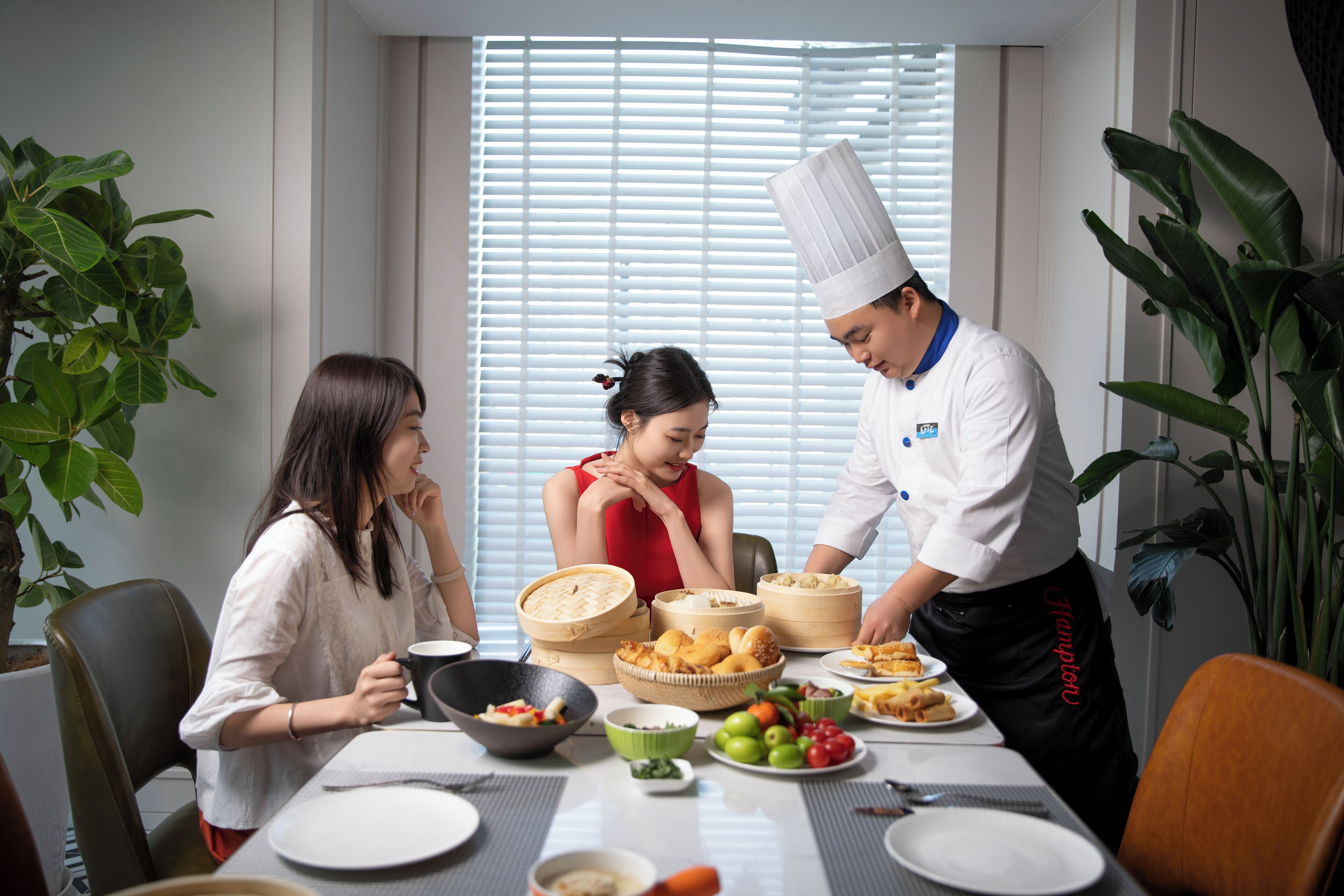A dining table with various dishes, a chef serving food, and two diners.