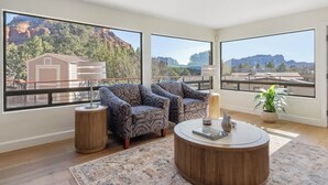 Une pièce spacieuse avec de grandes fenêtres donnant sur les montagnes et un bâtiment. On y trouve deux fauteuils à motifs, une table basse ronde et une petite table d’appoint.