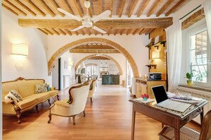Un vaste espace de vie doté d’un plafond en bois, d’un canapé, d’une table à manger avec un ordinateur portable, et d’une porte en arc donnant sur une cuisine.