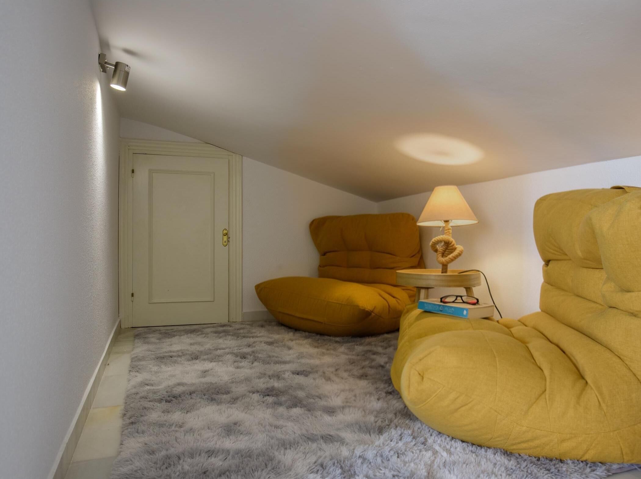 Een kamer met een deur, een hoekbank, een lamp en een klein tafeltje met boeken en brillen.