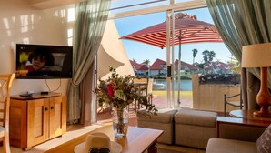 Ein Wohnzimmer mit Fernseher, einem Tisch mit Hut und Sonnenbrille, einer Couch und Blick auf einen Pool und Häuser.