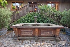 A rustic stone fountain with wooden plaques reading 'La Cascina' and '1890' in front of a wooden building with a wooden staircase and a lantern. - La Buona Stella Napa Valley (Saint Helena)