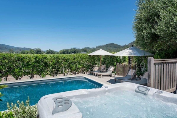 A hot tub with a pool, surrounded by lounge chairs and umbrellas, with a view of a vineyard and hills in the background.