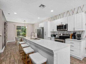 A modern kitchen with white cabinets, stainless steel appliances, a central island with bar stools, and a view to the outdoors. - Super Calm- Sleeps 17 W Designs Close TO Parks (Kissimmee)