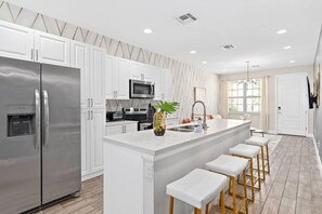 A modern kitchen with white cabinets, stainless steel appliances, a central island with bar stools, and a large window providing natural light. - Super Calm- Sleeps 17 W Designs Close TO Parks (Kissimmee)