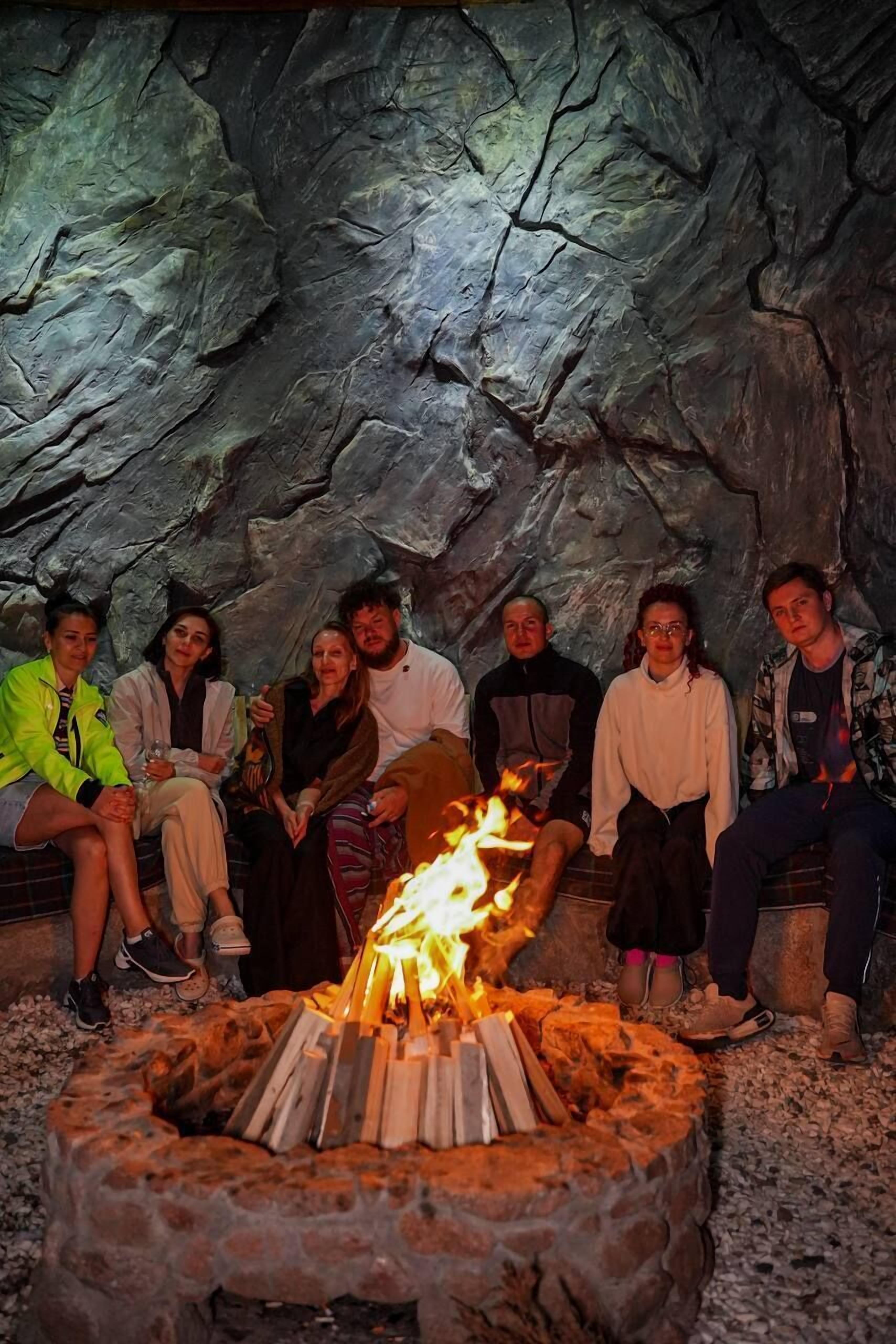 Eine Gruppe von Menschen sitzt um eine Feuerstelle aus Stein in einer höhlenartigen Umgebung.