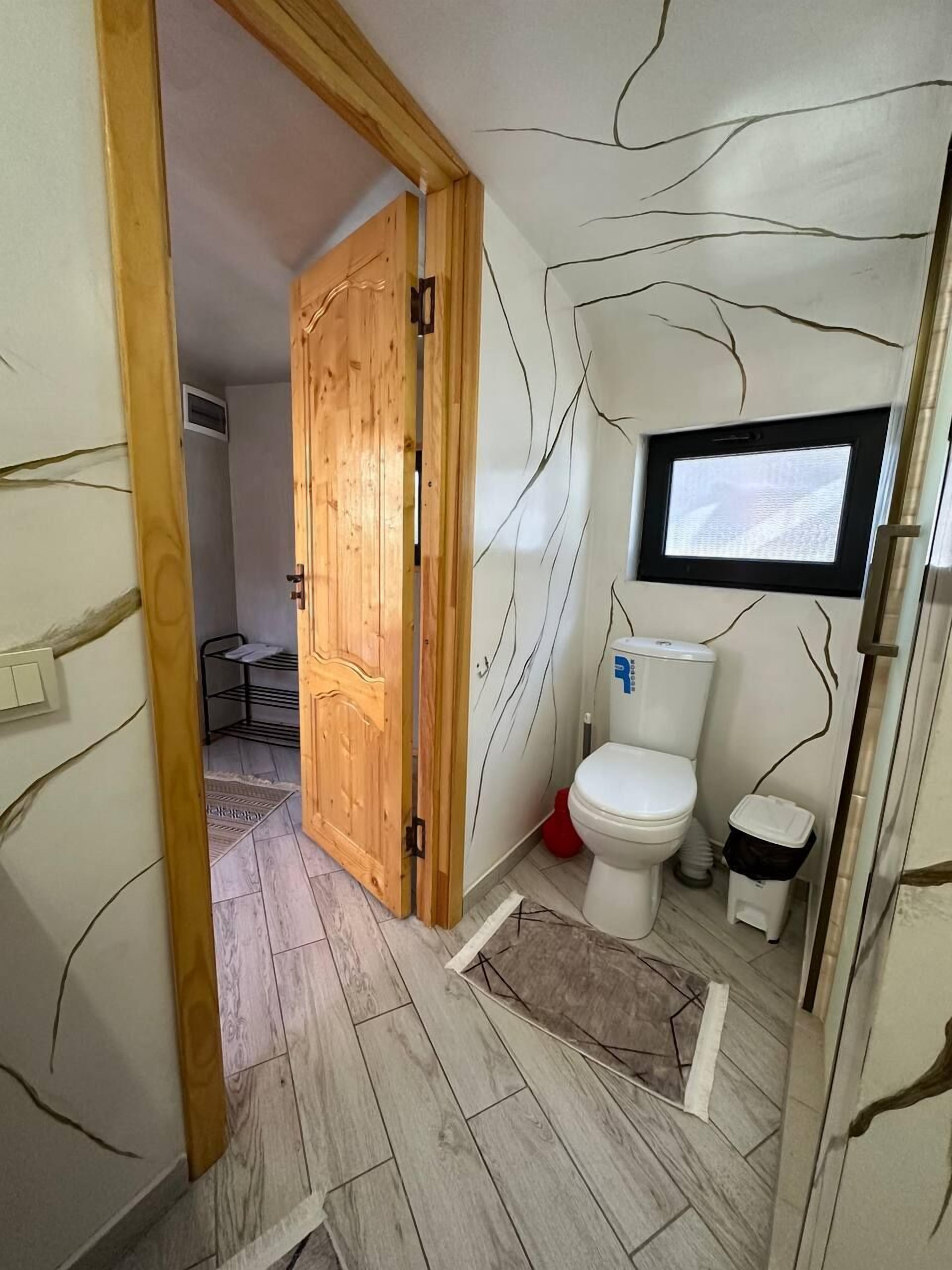 A compact bathroom with a wooden door, a toilet, a small trash bin, and a wall-mounted TV.