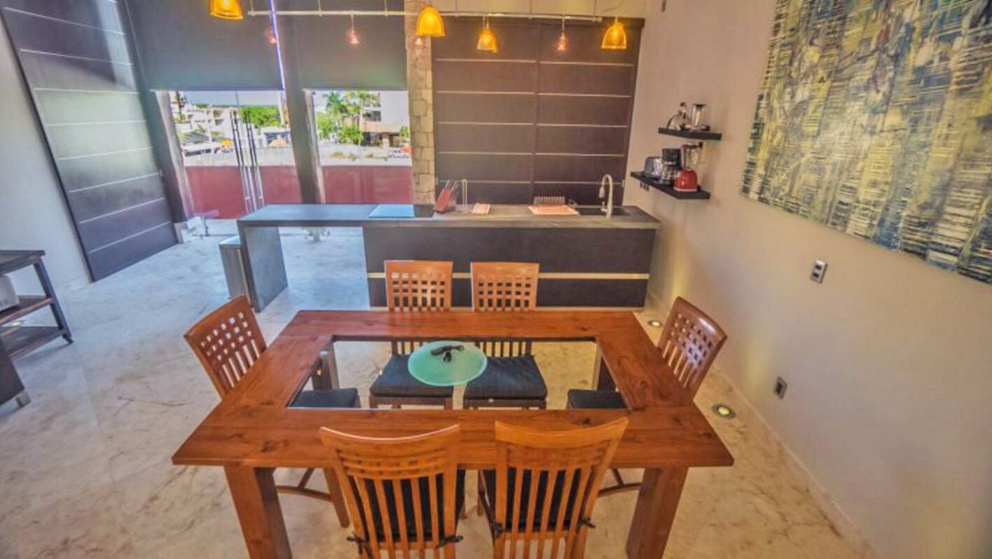 Comedor con mesa y sillas de madera, una encimera de cocina y vistas al exterior.