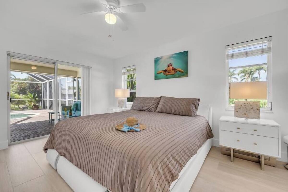 A bedroom with a bed, a ceiling fan, a painting, a nightstand, and a view of the outdoors.