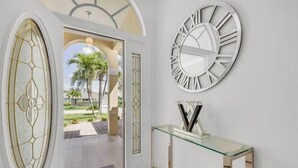 A modern interior with a glass console table, a large wall clock, and a door leading to an outdoor area with palm trees. - Villa Samba (Cape Coral)