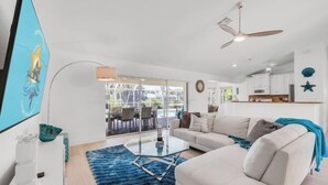 A modern living room with a white sofa, a glass coffee table, and a blue rug. There is a ceiling fan, a TV mounted on the wall, and a kitchen area visible through an open door. - Villa Samba (Cape Coral)