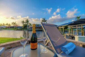 A table with a bottle of champagne and two glasses, a rolled towel, and a chair with a sunshade, overlooking a body of water and palm trees. - Villa Barefoot Cove (Cape Coral)
