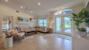 A spacious living room with a large sofa, a coffee table, and a dining area. There is a large window providing natural light and a view of the outdoors. - Villa Barefoot Cove (Cape Coral)