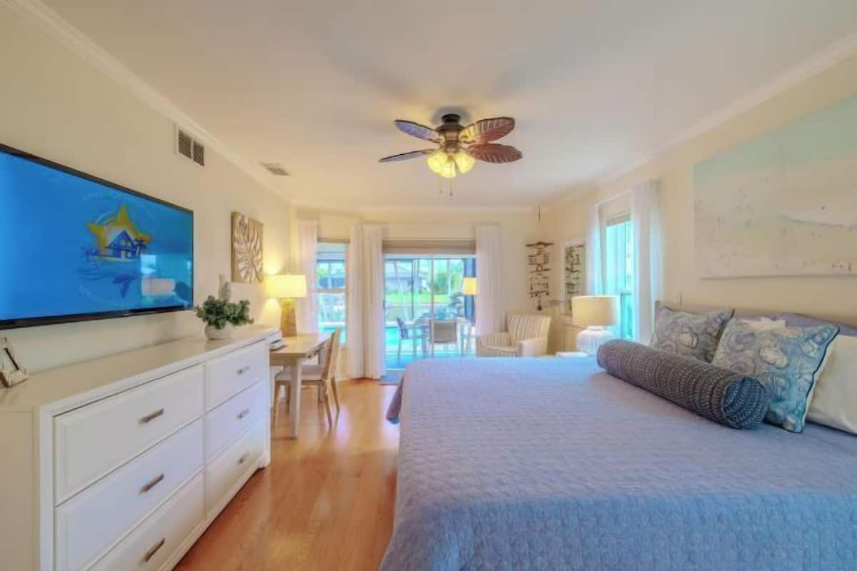 A bedroom with a bed, a TV, a ceiling fan, and a view of the outdoors.