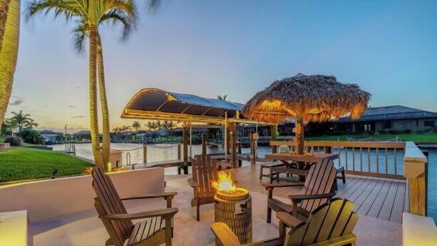 A deck with wooden chairs and a fire pit, overlooking a body of water and palm trees. - Villa Barefoot Cove (Cape Coral)